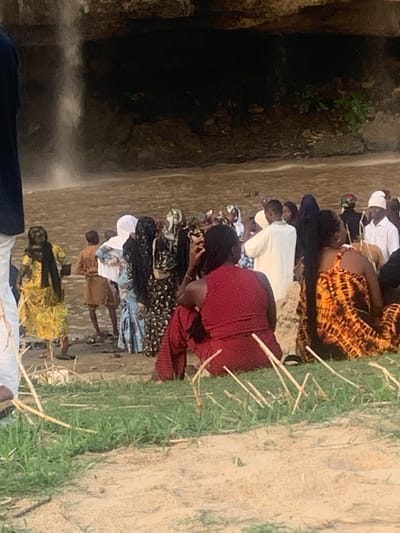 The image depicts a gathering of people at the Gurara Falls in Kukum, Nigeria. A line of approximately 10 individuals, mostly women, are positioned near the edge of the falls. They are kneeling or crouching, possibly in prayer or observation. The environment shows the rocky terrain and vegetation surrounding the waterfall. The waterfall itself is partially visible. People are wearing various attires, some with head coverings. Sticks are laid on the ground next to the kneeling persons.