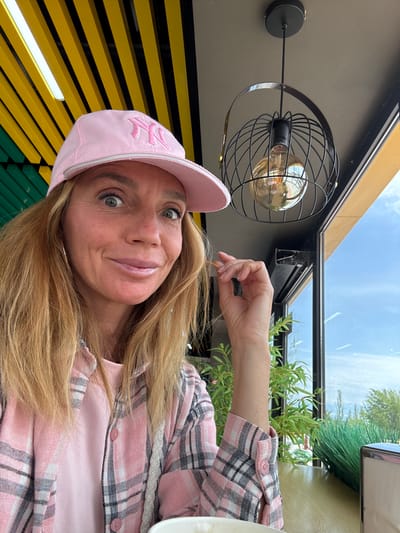 A woman with long blonde hair, wearing a pink baseball cap with a stylized "NY" or "YK" logo, and a pink plaid shirt over a matching pink t-shirt, smiles slightly at the camera. She appears to be seated outdoors or in a semi-outdoor setting, possibly a cafe or restaurant, in Кашат, Kyrgyzstan. Her right hand is raised near her head, lightly touching her hair. In the background, a modern light fixture with a black wire cage and an Edison bulb hangs. Above, a clear sky with scattered clouds is visible through what looks like a glass roof or canopy. To her left, vibrant yellow and black horizontal slat-like structures are visible, transitioning to green slats further down. Lush green foliage is visible through a glass partition behind her, suggesting an open-air or well-lit environment during the daytime.