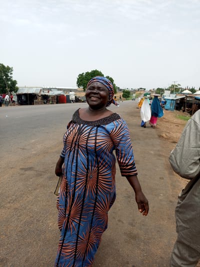 An adult Nigerian woman is pictured standing on a paved road in Gadao, Nigeria, with a light-colored, overcast sky above. She is smiling widely and looking towards the camera. She is wearing a blue and orange patterned dress with a matching headscarf. Her left hand is extended slightly, and she is carrying a bag. In the background, there are several open-air market stalls and buildings lining the road. Other people are visible in the distance, some walking and others gathered around the stalls. The road appears to be a main thoroughfare with vehicles present.