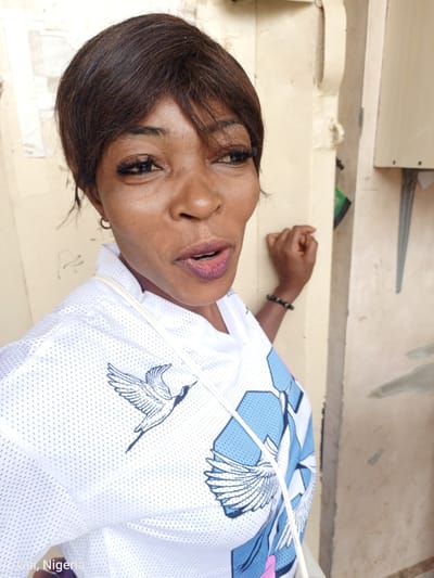 A medium shot captures a Black woman with dark brown hair and makeup standing in front of a weathered yellow wall in Kuje, Nigeria. She is wearing a white short-sleeved shirt adorned with a blue and white bird design, and a white strap from a bag is visible across her shoulder. She also wears a beaded bracelet on her left wrist. Her mouth is open, and she appears to be speaking or in mid-conversation. The background consists of the textured yellow wall and part of a gray concrete structure.