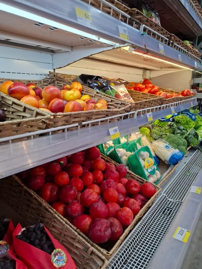 This image depicts a grocery store aisle, likely in Slyudyanka, Russia, showcasing a variety of fresh produce displayed on tiered shelving. The shelves are constructed of metal and wood, with white wire grids forming the base of each display.
In the foreground, a woven basket is filled with ripe, red plums. To the left of the plums, a red-ribboned container holds a cluster of dark purple grapes.
The middle shelf displays a variety of vegetables. Green plastic containers hold mushrooms. Behind the mushrooms are bunches of broccoli and heads of lettuce. A price tag labeled "219" is visible on the shelf edge.
The top shelf features a woven basket overflowing with peaches and nectarines, a mix of red and yellow hues. Further back, a basket is filled with bright red tomatoes, and next to them, other produce, possibly more tomatoes or other fruits, are visible. Price tags with numbers like "64" and "99" are affixed to the shelf edges.
The overall environment suggests a well-stocked grocery store with a focus on fresh produce. The lighting appears to be from overhead fluorescent fixtures, illuminating the colorful fruits and vegetables.