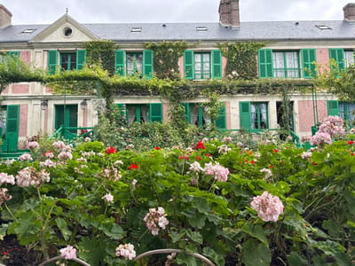 The image displays the front facade of a two-story residence in Giverny, France, identifiable as Claude Monet's house. The building features a light pink-peach stucco exterior with pale cream or beige trim, a dark gray slate roof with three visible dormer windows and two brick chimneys. All windows are fitted with open, bright green louvered shutters. Extensive green ivy and other climbing plants cover significant portions of the facade, especially between windows and along the upper story. A green-painted wooden pergola, supporting climbing vegetation, spans horizontally across the front of the house above the ground floor. Two dark, wall-mounted lanterns are positioned near a ground-floor entrance, which is fronted by a small green wooden railing. The lower half of the image is dominated by a dense garden bed containing numerous large-leafed plants, predominantly bearing clusters of light pink flowers and scattered vibrant red flowers. No individuals or specific actions are depicted. The sky is overcast and gray.