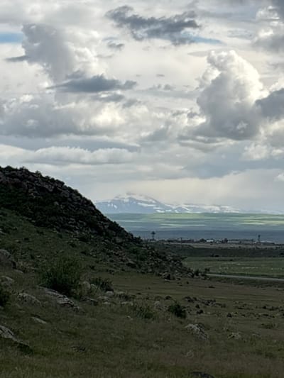 The image shows a landscape in the vicinity of Tlik, Armenia. The foreground features a rocky, grassy hillside, sloping upwards from right to left. In the midground, there is a view of cultivated fields and distant buildings or structures under a sky with prominent clouds. There is no visible human presence or interaction in the scene.