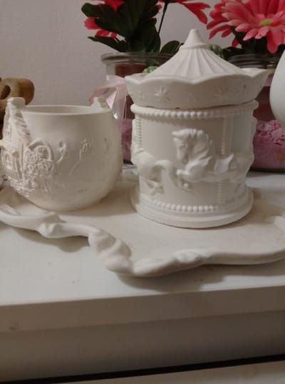 The image shows two decorative ceramic items on a white surface. On the left is a mug with embossed detailing of a house and figures. To the right is a carousel-themed jar with horses and a ridged lid. Behind these items, blurred pink flowers in a glass container are visible. The overall scene is a still life of decorative objects, possibly on a shelf or table. The location in Wedel, Germany is not discernible from the image.