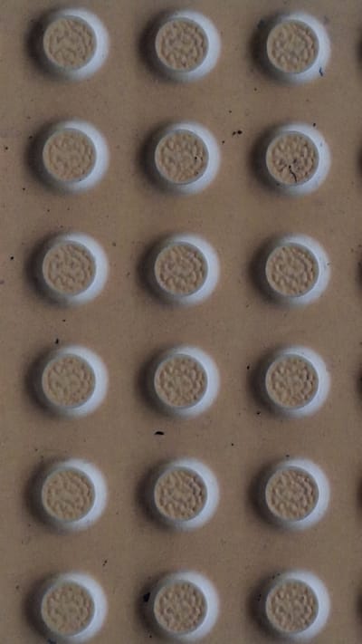 The image shows a close-up of a tactile paving surface, typically used to aid visually impaired pedestrians. The surface consists of rows and columns of raised, circular bumps, each featuring a textured top surface. The bumps are lighter in color than the surrounding flat surface, which appears to be a beige or light brown color. There are some scattered dark spots or debris on the flat surface. There is no identifiable location in Karawang, Indonesia, or any evidence of people, objects, actions, or interactions in the image.