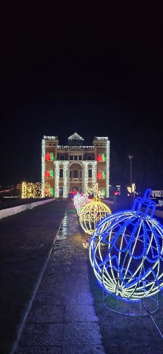 A building, decorated with red, green, and white lights, stands beside large glowing spherical structures. The lights are strung along the building's facade, highlighting its architectural details. The spherical lights, one blue and one yellow, add to the festive atmosphere. The scene is set at night, with darkness surrounding the brightly lit structures. A walkway can be seen in the foreground, reflecting some of the lights.