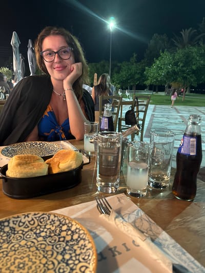 A young woman with dark hair and glasses is seated at a table outdoors at night. She is wearing a black jacket over a blue and yellow patterned dress. Her right hand is resting on her chin, and she is looking towards the camera with a slight smile. On the table in front of her are plates, glasses, bottles of soda, and bread rolls in a black dish. The background shows trees and other diners. The scene appears to be a restaurant in Asprovalta, Greece.