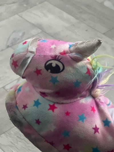 The scene is located in the city of Пугачёво, Russia. The primary subject is a plush, unicorn-themed slipper or toy, depicted from an elevated perspective. The object is multicolored in pastel pink, blue, and light yellow, featuring a scattered pattern of small hot pink and light blue stars. A large, stylized dark eye with an eyelash design is visible on the left side of the head. A silver-colored, conical horn protrudes from the forehead, displaying minor wear at its tip. Behind the horn, several tufts of pastel-colored fabric, including yellow, light green, and purple, simulate a mane. A light-colored fabric strap or band spans horizontally across the top of the head, appearing torn or detached on its left side. The plush material shows signs of general use, and a dark scuff mark or discoloration is present on the lower left edge of the object. The object rests on a light grey or off-white floor surface, which exhibits a subtle, irregular pattern suggestive of tile or wood grain. No individuals or active interactions are present in the frame.