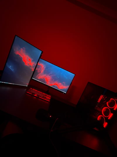 The image shows a gaming setup against a red wall. There are two monitors displaying a nebula-like image with red and blue tones. A red backlit keyboard is visible on the desk. A computer tower with red LED fans is positioned above the monitors. A mouse and mousepad are dimly visible to the right of the keyboard. The location is not discernible from the image.