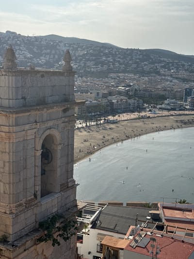 The image, taken in Peníscola, Spain, captures a vibrant coastal scene encompassing historical architecture, a lively beach, and a mountainous cityscape under clear skies. In the immediate foreground, occupying the lower right side, stands a robust stone bell tower or part of a historic stone building, likely a church or castle. Its weathered blocks and arched openings, with small patches of greenery growing on the masonry, suggest significant age.
Beyond this ancient structure, a wide sandy beach unfolds, bustling with numerous people. Many individuals are visible sunbathing on loungers, relaxing under beach umbrellas, or strolling along the shore. A paved promenade, lined with scattered trees, separates the sand from a row of modern buildings, characteristic of a developed coastal town.
The calm, light-blue sea stretches into the distance, where several people can be seen swimming or engaged in water sports like paddleboarding or kayaking. The background is dominated by a majestic hill or mountain, its slopes densely covered with white and light-colored buildings, indicating extensive urban development that cascades down towards the sea. The sky is bright and clear, suggesting a sunny day, likely morning or early afternoon, perfect for outdoor and beach activities. The overall atmosphere is one of relaxation and leisure. On a building in the far lower right, barely visible, the word "VISTA" is printed, reinforcing the scenic nature of the location.