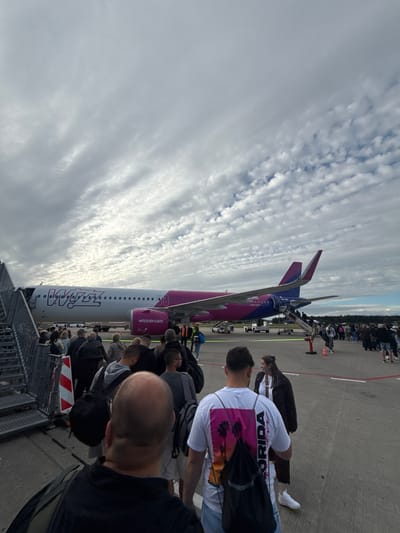 This image depicts a group of passengers boarding a Wizz Air airplane on a tarmac. The airplane, a pink and purple Airbus A320, is parked with its stairs extended. Passengers are queuing to board, some with carry-on luggage. In the background, other passengers and ground crew are visible, as well as a line of trees and buildings, suggesting an airport environment. The sky above is cloudy. The location is identified as Künersberg, Germany. The image captures a moment of travel, with the focus on the boarding process of an airline flight.