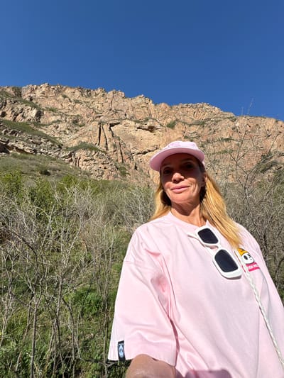 A woman with long blonde hair smiles softly at the camera in an outdoor setting, likely a mountainous or hilly landscape. She is wearing a pink baseball cap and a pink long-sleeved shirt. White rectangular sunglasses are perched on the front of her shirt.
The background is dominated by a towering, rocky mountain face under a clear, deep blue sky. Sparse, dry vegetation, including bare branches and some green bushes, is visible in the mid-ground and foreground. The bright sunlight suggests it is daytime.
Notable details include a partially visible design on her pink cap (possibly stylized letters) and an embroidered patch on her shirt near her right shoulder (viewer's left) featuring a cartoon character, possibly a duck, with the partially legible text "TOR H..." below it. A small, dark, circular logo is also visible on the cuff of her left sleeve (viewer's right). The scene depicts a rugged natural environment, consistent with the Cholpon Ata, Kyrgyzstan location context.