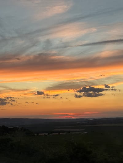 This is a vertical, low-angle photograph of a sunset over a rural landscape. The sky is filled with dramatic clouds, painted in hues of orange, pink, and pale blue, transitioning into a deep orange glow near the horizon. wispy clouds are scattered throughout, some appearing darker and more defined against the lighter sky. The overall impression is one of serene beauty and natural spectacle.

Below the sky, a dark, silhouetted landscape unfolds. The terrain appears hilly or mountainous, with a distinct line of trees or vegetation forming a dark band against the lighter sky. In the distance, faint structures, possibly buildings or farmland, are visible. The lower right portion of the image is dominated by dark green, suggesting fields or dense vegetation, also silhouetted against the fading light.

The time of day is clearly sunset, given the warm, saturated colors in the sky and the diminishing light on the landscape. The weather appears calm, with the clouds suggesting a clear or partly cloudy sky that is being illuminated by the setting sun. The location appears to be a rural area, likely in a region with rolling hills or mountains, as indicated by the terrain and the presence of open fields. No people or specific objects are clearly discernible in the image. The image evokes a sense of tranquility and the awe-inspiring beauty of nature. There is no visible text in the image.