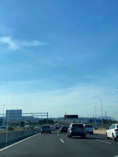 The image depicts a traffic jam on a highway in Sant Joan Despí, Spain. A line of vehicles is visible, including cars, vans, and possibly trucks, all stationary or moving very slowly. The sky is blue with some clouds. Overhead signs and highway lighting are visible alongside the road. Some of the vehicles' license plates are visible. In the background, there's a landscape with hills and possibly some industrial or commercial structures.