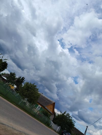 The image shows a tilted view of a rural scene under a dramatic, cloudy sky. In the foreground, a paved road runs diagonally from the bottom left to the upper left. A green metal fence runs parallel to the road. Behind the fence, a yellow house with a grey roof is visible, featuring green shutters on a window. Trees with dense green foliage are scattered throughout the scene, some behind the fence and some further back. In the upper portion of the image, a dark bird is seen flying in the sky. The sky is filled with a mix of dark, heavy clouds and patches of blue sky with lighter clouds. The overall impression is of a potentially stormy or overcast day in a rural Belarusian village.