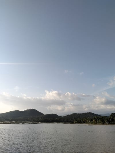 The image depicts a serene landscape. Still water fills the foreground, reflecting the sky above. Behind the water, low, rolling hills covered in lush greenery stretch across the horizon. A clear blue sky dominates the upper portion of the image, punctuated by scattered, soft white clouds. The scene exudes tranquility and peace.