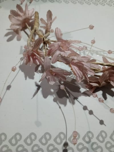 A delicate arrangement of blush-pink fabric flowers and beaded sprigs rests on a patterned surface. The flowers feature narrow, textured petals clustered around a central stem, while the sprigs are adorned with small, spherical beads along slender, transparent strands. The arrangement casts soft shadows on the background, revealing subtle details in the fabric and bead textures. The patterned surface is a white with a gray letter "S" pattern repeating diagonally.