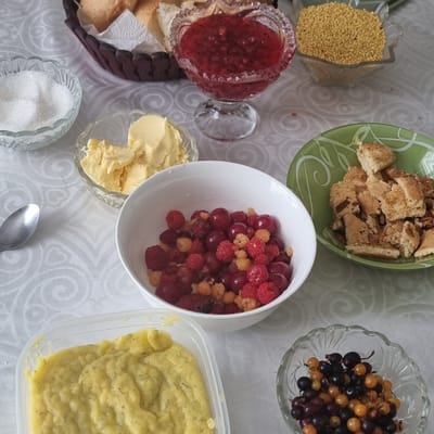 The image depicts a table set with various food items, suggesting a meal or gathering. On the table, there is a basket of bread, a bowl of red jam, a bowl of small yellow grains, a bowl of white sugar, a bowl of butter, a white bowl filled with red and yellow berries, a green plate with broken pieces of pastry, a clear plastic container with a yellow spread, and a small glass bowl containing black and yellow berries. The background is a white tablecloth with a subtle swirling pattern. There are no people visible in the image. The lighting appears to be natural, coming from above and to the right. The location is described as Ақкемер, Kazakhstan.