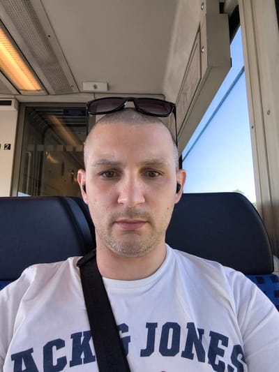 The image shows a close-up selfie of a man sitting inside a train, likely during daytime as indicated by the bright sky visible through the window and the interior lighting. The man has a shaved head and is wearing a white t-shirt with the text "JACK JONES" printed on it in dark blue letters. He also has dark sunglasses resting on top of his head and is wearing black wireless earbuds. The background reveals the interior of a train carriage with dark blue seats and a window showing a clear blue sky. The overall mood appears casual and relaxed, possibly during a commute or a journey. There is no indication of specific weather conditions beyond the clear sky. The man is looking directly at the camera, and his expression is neutral. The location context is Koserow, Germany.
