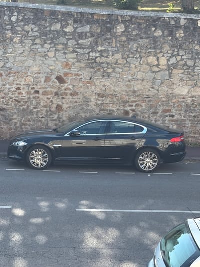 A black Jaguar sedan is parked on a street in Limoges, France, beside a stone wall. The car is viewed from the side, with its left side facing the camera. The vehicle has a sleek, modern design with chrome accents and alloy wheels. The stone wall behind the car is constructed from irregular-shaped stones of varying sizes and colors, creating a rustic texture. The street is paved with asphalt and marked with white lane dividers. Sunlight casts dappled shadows on the road surface, suggesting it is daytime. A portion of another vehicle, possibly a silver hatchback, is visible in the lower right corner of the frame, suggesting a typically urban environment.