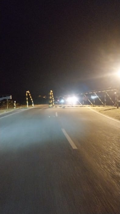The image captures a nighttime scene on an asphalt road in Keudearon, Indonesia. The road surface exhibits reflections of artificial light, suggesting a wet or highly reflective texture. A solid white lane marking is visible on the left side of the road. In the mid-ground, two distinct vertical bollards, striped in black and yellow/white, frame what appears to be an entrance or checkpoint. Further down the road, a horizontal barrier or gate, similarly striped or reflective, blocks the thoroughfare. Intense glare from a bright light source, possibly a streetlamp, emanates from the upper right. Directly ahead, beyond the gate, strong headlights from an unidentifiable vehicle are visible, producing significant glare and partially obscuring the vehicle. The sky above is dark. No individuals are present in the visible frame.