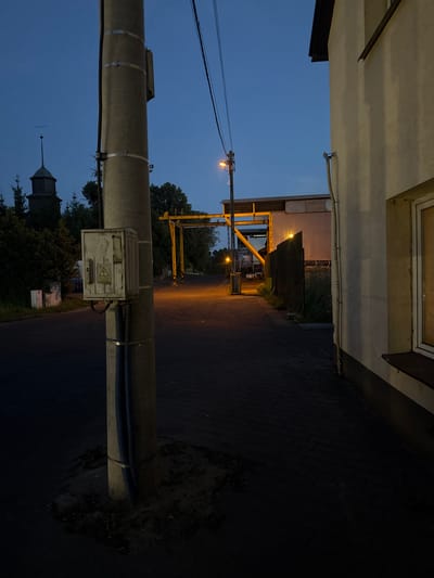 In Wąwelnica, Poland, at dusk, a concrete utility pole stands prominently in the foreground on the left side of a paved pathway. Attached to the pole is a light-colored electrical junction box with a warning sticker displaying a lightning bolt symbol. Dark blue conduits run vertically along the pole, secured with metal bands. Beyond the pole, the pathway extends into the distance, illuminated by a single streetlamp casting an orange glow. To the left of the pathway, a dark wooden church tower with a cross on its spire is visible amidst trees, and further back, a large industrial building with a yellow gantry structure is situated. On the right side of the image, a light-colored building with a window and a dark overhang is partially visible. The sky is a deep blue, indicating the late hour of the day. The environment appears to be a semi-urban or industrial area.