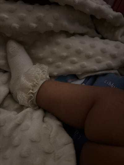 The image shows a close-up of a child's leg wearing a white sock with a ruffled trim. The leg is resting on a soft, white blanket with a dotted texture. In the background, there is a glimpse of a blue fabric with white lettering, possibly a diaper or clothing item, with the word "Libb" partially visible. The scene is dimly lit, suggesting an indoor setting. The location of Geulanggang Teungoh, Indonesia, is not visually evident in this image.