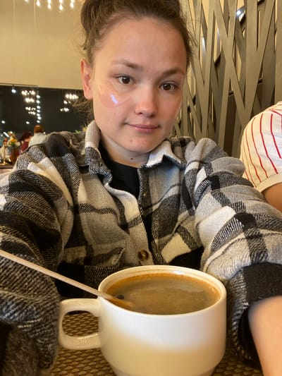 A person, possibly female, with light skin and brown hair is seated inside a likely restaurant or cafe. The person is wearing a black and white flannel over a striped red and white shirt. A white mug with a beverage is to the person's right, along with a wooden coffee stirrer. There is a clear under eye patch on the subject's left under eye. Behind the person, blurred, is a dark window with decorative lights, and a glimpse of what appears to be additional tables, chairs, and a decorative flower arrangement. The image has been taken at a rotated angle. There is no information to support the location in the city of Peresvet, Russia.