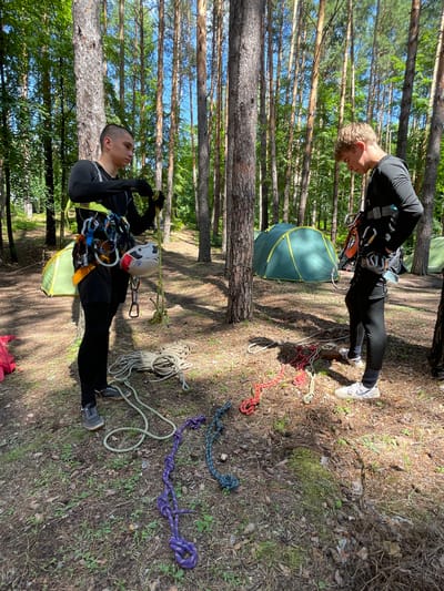 Two young men, likely teenagers, are standing in a sun-dappled forest clearing, surrounded by tall pine trees. The ground is covered with pine needles, fallen leaves, and sparse green vegetation. In the background, two green tents are pitched.
Both individuals are dressed in athletic wear suitable for outdoor activities. The man on the left wears a black long-sleeved shirt, black shorts over black leggings, and dark athletic shoes. He has a harness with carabiners and other climbing gear around his waist and is actively manipulating a rope or line with his gloved hands. A white and red climbing helmet is attached to his harness.
The man on the right wears a black long-sleeved shirt, black leggings, and white athletic shoes. He also has a climbing harness on and appears to be looking down at the ropes on the ground.
Scattered on the ground between the two men are several coiled ropes of various colors, including white, red, purple, and blue. Some ropes are neatly coiled, while others are more loosely arranged. The overall scene suggests an outdoor recreation or training activity related to climbing or ropes courses. The location is identified as the village of Tiapino, Russia, based on the provided metadata.