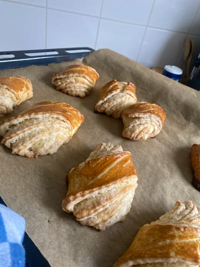 The image displays eight golden-brown, crescent-shaped pastries with visible laminated dough structure, resting on a light brown parchment paper-lined baking sheet, located in a kitchen in Chantepie, France. An additional pastry, darker in appearance, is partially visible on the far right edge. The pastries are irregularly spaced across the parchment paper. In the background, a white tiled wall with square tiles is present. A dark grey metal oven rack is partially visible in the upper-left background. To the upper right, a white cylindrical container with a blue lid and dark kitchen utensils are partially visible against the tiled wall. A blue and white checkered fabric item is visible in the lower-left foreground.