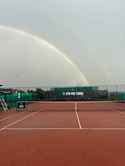 The image displays an outdoor red clay tennis court located in Giebułtów, Poland, under an overcast, light-grey sky. A distinct double rainbow is visible arcing across the upper portion of the sky. The tennis court is marked with white lines and features a black net with a white center strap positioned at its midpoint. Behind the net, a large black banner is stretched horizontally, bearing the white text "UTR PRO TENNIS TOUR" and its logo. Additional green wind screens, some featuring "HEAD" logos, line the perimeter fencing of the court. To the left side of the court, a smaller black banner displaying "UTR PRO TENNIS TOUR" stands near a white metal umpire chair. No individuals are present on the court or in the immediate vicinity.