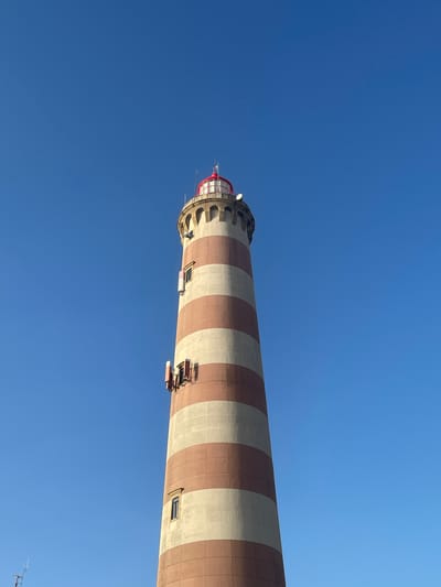 The image depicts the Barra Lighthouse in Gafanha da Nazaré, Portugal, under a clear, vibrant blue sky. The lighthouse is a tall, cylindrical structure with alternating horizontal bands of light beige and reddish-brown. It has a red-capped lantern room at the very top, surrounded by arched windows and a railing. Several small rectangular windows are visible along the body of the lighthouse, and on one side, there are multiple antenna-like structures attached to the exterior. The perspective is looking up at the lighthouse from a low angle, emphasizing its height against the vast expanse of the sky. There are no people or animals visible in the frame. The environment appears to be an open area, typical of a coastal location where a lighthouse would be situated.