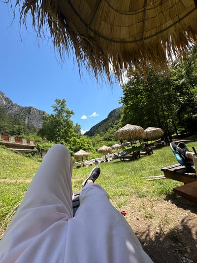 The image depicts a sunny outdoor scene, likely in Zgorigrad, Bulgaria, with a person's legs visible in the foreground, wearing light-colored trousers and black sneakers. They are seated on a wooden bench, possibly at an outdoor seating area. In the background, there are several thatched umbrellas providing shade over tables and benches where other people are seated. The environment is lush and green, with a clear blue sky overhead. Mountains and dense trees form the backdrop, and a small bridge with red railings is visible to the left. The overall atmosphere suggests a relaxed, recreational setting, possibly a park or a riverside cafe.