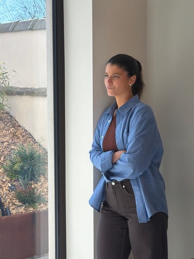 The scene is set indoors, within the city of Marche-lez-Écaussinnes, Belgium. A female subject is positioned in profile, standing near a large window with a black frame, leaning against a light-colored interior wall. Her dark hair is pulled back into a ponytail, and she wears light-colored stud earrings. She is dressed in a brown tank top beneath an unbuttoned light blue, plaid-patterned, long-sleeved shirt, and dark brown trousers. Her arms are crossed over her chest, and she looks outward through the window with a slight smile. The view through the window reveals an exterior area under a clear blue sky, featuring a light beige stucco wall, a ground covered with wood chips, and various green plants, including bamboo-like foliage and a bush, partially contained within a visible dark brown planter box.