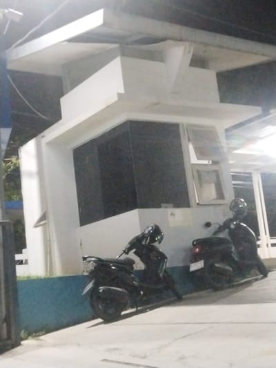 In the city of Lampeuneurutblang, Indonesia, two dark-colored motorcycles are parked outside a white, multi-level structure at night. The structure has a prominent dark-paneled section and windows. One motorcycle is positioned in the foreground to the left, and the other is further back to the right, partially obscured by the first. The lighting suggests an outdoor setting, possibly a parking area or a service station.