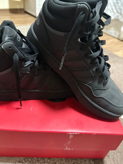 The scene is an indoor environment in Novgrad, Bulgaria. Two black high-top athletic shoes are prominently displayed, positioned on top of a red rectangular shoebox. Both shoes feature black laces, black midsoles, and the characteristic three stripes on their sides, consistent with Adidas branding visible on parts of the shoes. The left shoe has an untied black lace hanging vertically towards the box surface. The right shoe has its laces tied. The red shoebox exhibits several small white scuff marks or residue stains on its top and upper front surfaces. The shoes and box are resting on a light brown, textured carpet or rug. The background is out of focus, showing a light-colored piece of furniture, possibly a dresser or cabinet, against a light-colored wall. No individuals are present, and no active actions are depicted.