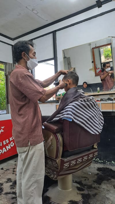 In a barber shop located in Kruengjuli Timu, Indonesia, a male barber, wearing a white face mask and a brown patterned shirt, is in the process of cutting the hair of a young boy. The barber holds a comb in his left hand and an electric hair clipper in his right hand, actively working on the boy's head. The boy is seated in a vintage barber chair, draped with a striped cape. In the background, a mirror reflects another man, also wearing a face mask, who appears to be the barber in an adjacent station, and a woman wearing a headscarf. The floor around the barber chair is scattered with cut hair, indicating recent activity. The environment appears to be a simple, utilitarian establishment with wooden structural elements and a white painted ceiling.