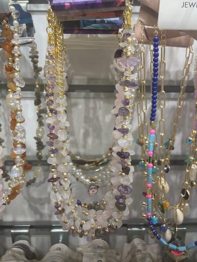 The image shows a close-up of several necklaces displayed on a rack. The necklaces are made of various beads, including polished stones in shades of pink, purple, white, and orange, as well as pearls and small gold-colored spherical beads. Some of the necklaces also feature charms, such as small shells and a blue evil eye. The background appears to be a retail environment, with shelves and the reflection of lights. The overall impression is of a jewelry display. This image was captured in Yunus Emre, Turkey.