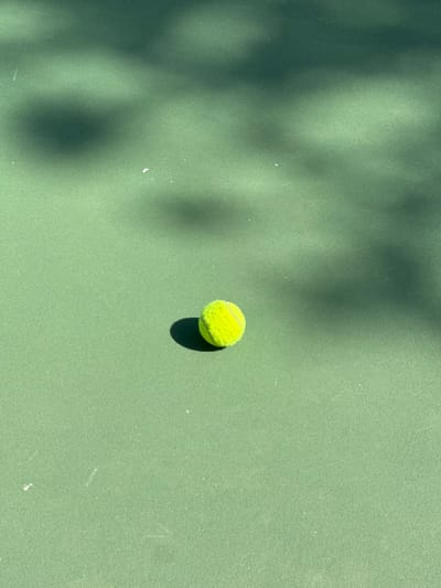 A single fluorescent yellow-green tennis ball rests on a light green, textured hard court surface, consistent with a tennis court, in the city of Ericeira, Portugal. The ball exhibits visible felt nap. Direct sunlight illuminates the scene, casting a distinct dark shadow directly behind and to the left of the ball. Additional diffuse, irregular dark shadows, indicative of overhead foliage or structures, are present across the upper left and central-upper sections of the court surface. Small white flecks are scattered on the court. No individuals or active interactions are depicted.