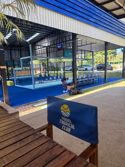 This image captures an outdoor padel tennis court facility in Ban Bang Rak, Thailand, likely during daylight hours as indicated by the natural lighting and shadows. The foreground features a weathered wooden bench or table, with a blue fabric banner displaying the text "PADEL TROPICAL CLUB" and a graphic of a tennis ball.
The central focus is a vibrant blue artificial turf padel court enclosed by glass walls and metal fencing. A light blue metal frame surrounds a section of the court. To the left of the court, a woman wearing dark athletic wear is bent over, tending to shoes and a black trash bag. Several yellow padel balls are stacked in a corner near the court. A row of blue stadium-style chairs is positioned on a raised platform behind the court.
In the background, parked vehicles, including a white SUV and a black SUV, are visible on a paved area beyond the court. Lush green trees and vegetation surround the facility, and a building with a corrugated metal roof and blue trim is visible above the court structure. A digital scoreboard displaying "8:43" is mounted on the wall of the building. The overall impression is of a well-maintained sports facility set in a tropical environment.