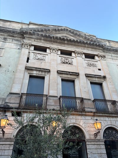 The image shows a section of a building in Acireale, Italy. The facade is made of light-colored stone with ornate details, including decorative carvings around the windows. The windows are rectangular, except for the ones on the right, which are arched. Each window has dark glass and a wrought-iron balcony. A street lamp is mounted on the building between the arched windows. A tree with dense foliage is partially obscuring the lower right of the image. The sky is visible in the upper left corner.