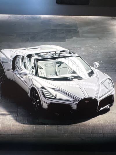 The image shows a close-up, top-down view of a white sports car with black racing stripes, possibly a Bugatti, parked on a textured, gray surface that appears to be a street or pavement. The car has a sleek, aerodynamic design with a prominent black rear wing or spoiler. Reflections of the surroundings are visible on the car's glossy surface, suggesting it's outdoors. The background is a dark, textured wall, possibly brick or concrete, with a grid pattern. The lighting suggests it could be daytime, with shadows cast on the ground. No people are visible in the frame. The scene is static, with the car stationary. The overall impression is one of a high-performance vehicle in an urban or industrial setting.