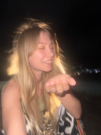 A young woman with blonde hair is leaning forward with her eyes closed and a smile on her face, blowing a kiss towards the camera. Her right hand is extended towards the viewer. She is wearing a green top and a patterned skirt. The background is dark, with blurry lights in the distance, suggesting a nighttime scene. The location is specified as Mare Anglaise, Seychelles.