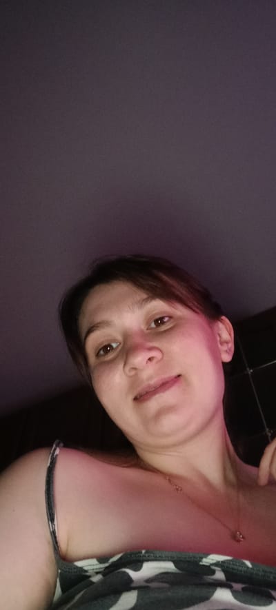 A young woman with brown hair and brown eyes is taking a selfie. She is wearing a camisole with a camouflage pattern. Her skin is flushed with pink from the lighting. She is wearing a delicate necklace with a small pendant. The background is dark and appears to be an indoor setting. The image is taken from a low angle, looking up at the woman. There is no identifiable information regarding the location in the city of Хохлы, Russia.