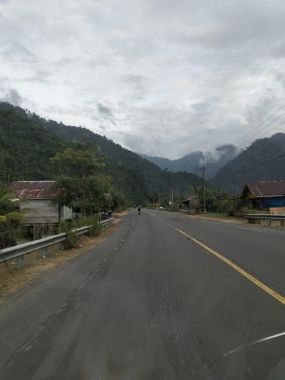 An outdoor, long-shot photograph depicts a two-lane asphalt road traversing a rural, mountainous landscape in Meunasahcot, Indonesia, under an overcast sky. The road, featuring a double solid yellow center line, extends into the distance, narrowing towards a valley between steep, heavily forested mountains. A metal guardrail runs along the left side of the road in the foreground. Beyond this guardrail on the left, a weathered wooden house on stilts with a rusty corrugated metal roof is visible amidst dense green foliage. Further down the road on the right, another structure with a similar roof is partially visible, along with a utility pole and overhead power lines. A single individual is observed riding a motorcycle away from the camera in the right lane of the road, approximately mid-distance. The mountains in the background are draped in thick jungle, with low-lying clouds or mist partially obscuring their upper slopes. The overall environment appears humid and undeveloped, with sparse human infrastructure integrated into the natural setting.