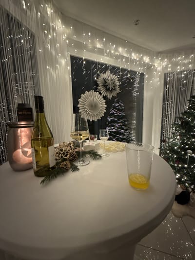 A festive table setting features a wine bottle in an ice bucket, a clear pitcher with lemon slices, wine glasses, and decorative greenery. The table is white, possibly covered with a tablecloth. A Christmas tree is visible in the background. Fairy lights adorn the window, with decorative paper fans. The scene conveys a celebratory, elegant, and cozy ambiance.