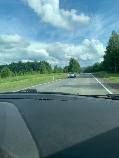 The image captures a view from inside a vehicle traveling on a two-lane asphalt road through a rural landscape. The road is lined with grassy verges and mature trees on either side. In the distance, a silver car is driving in the opposite direction. The sky is a bright blue with scattered white cumulus clouds, suggesting fair weather. The foreground shows the textured dashboard of the car, including a windshield wiper. The scene appears to be located in or around the city of Sudag, Russia, given the context of the request.