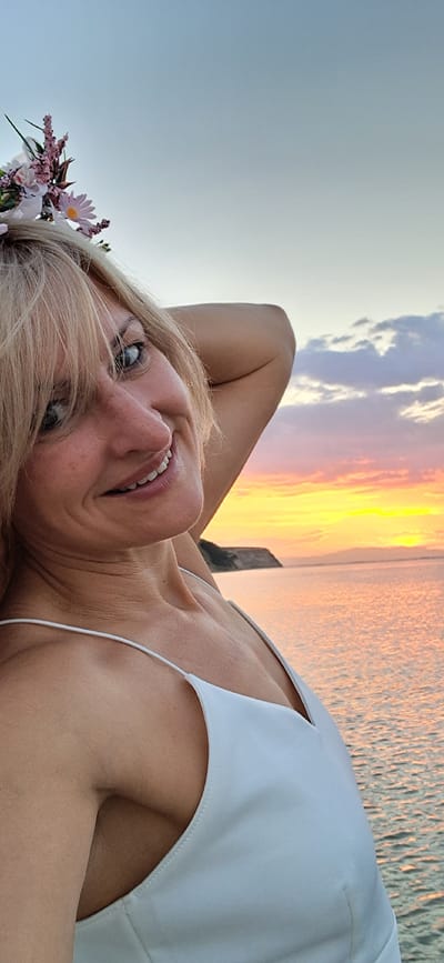 A blonde woman wearing a white dress and a flower crown poses with her hand behind her head against a sunset over the sea. The image is a selfie taken outdoors in Agia Triada, Greece. The sky is painted with hues of orange, pink, and yellow, with scattered clouds. The sea reflects the vibrant colors of the sunset. In the background, a faint silhouette of land is visible. The woman is smiling and looking towards the camera, her hair is short and styled with some strands falling over her face. The flower crown is adorned with small white and pink flowers. The white dress has thin spaghetti straps.