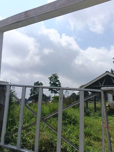 A landscape framed by a concrete structure and a metal fence. Lush green vegetation fills the midground, punctuated by tall trees. A structure with a peaked roof is partially visible on the right side of the frame. Above, the sky is filled with white clouds under a bright blue sky. The view is obstructed by the metal fence, partially obscuring the landscape.