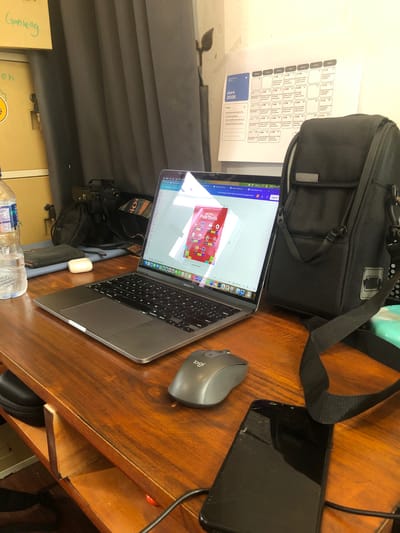 The image captures an indoor workspace from a top-down, slightly angled perspective, showcasing a desk cluttered with various items. The central subject is a silver MacBook Pro laptop, open and displaying a colorful digital schedule, likely related to the "Piala Dunia" (World Cup), with text "JADWAL Piala Dunia" prominently visible. To the right of the laptop is a grey Logitech mouse.
Behind the laptop, a black shoulder bag with various compartments is visible. Further to the right, a black smartphone with a visibly cracked screen lies flat on the wooden desk surface, connected by a black cable. In front of the laptop, a clear plastic water bottle, an AirPods case, and a camera peeking out from under the laptop are discernible.
To the left of the laptop, taped to a light-colored wall or partition, is a printed paper calendar for "Juni 2026". The calendar lists days of the week in Indonesian (Senin, Selasa, Rabu, Kamis, Jumat, Sabtu, Minggu) and outlines daily activities such as "Kumpul/Briefing Tim" (Team Meeting/Briefing), "Content/Copywriting", and "Planning Brand". On the laptop's top bar, the time "9:15" and date "Thu 4 Jun" are visible, along with a battery indicator at "37%". The word "Ganteng" and the number "५०" (50) are handwritten on a surface to the far left. The overall setting suggests a productive work environment, possibly in Cibinong, Indonesia, given the context and language.